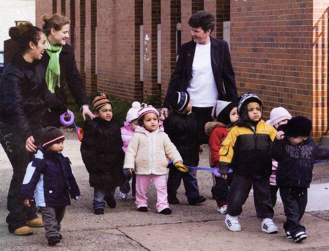 Three women smile at each other while walking with a group of small toddlers.