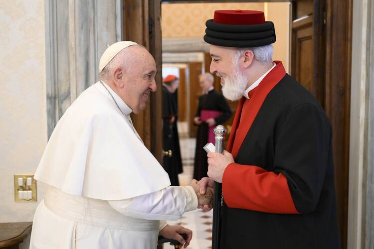 A man wearing white garments and a white hat shakes hands with a man wearing black and red garments