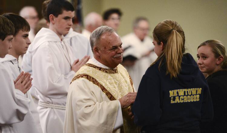 A priest in white robes speaks with a young girl in a jacket that reads "Newtown Lacrosse"