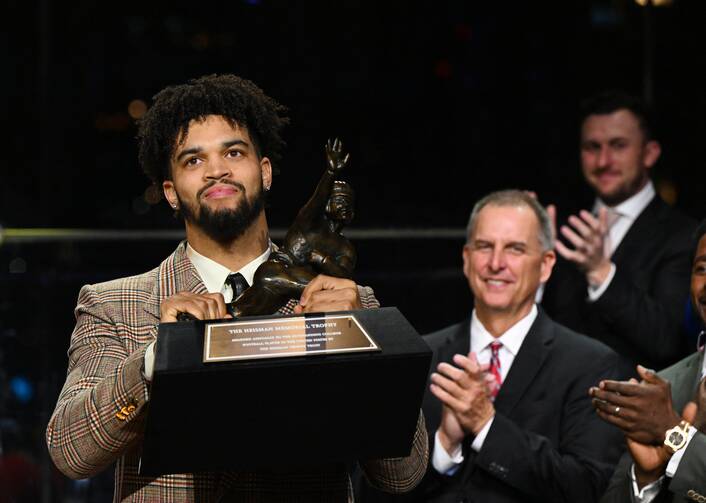 A man in a jacket and tie holds a football trophy while another man nearby claps