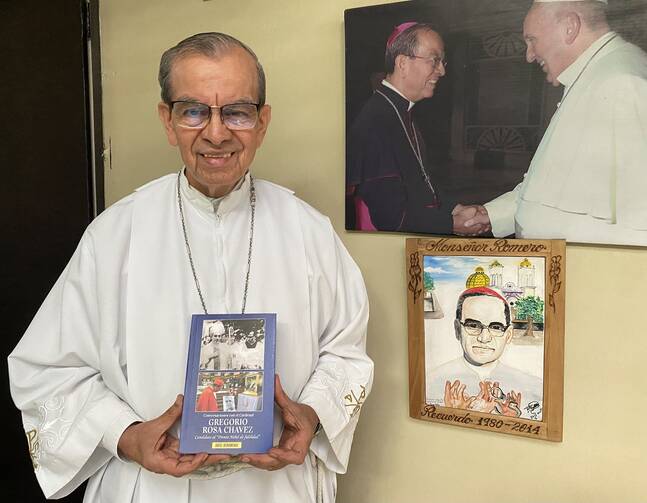 cardinal gregorio rosa chavez holds a copy of a book with a photo of oscar romero and a photo of oscar romero with the pope behind him