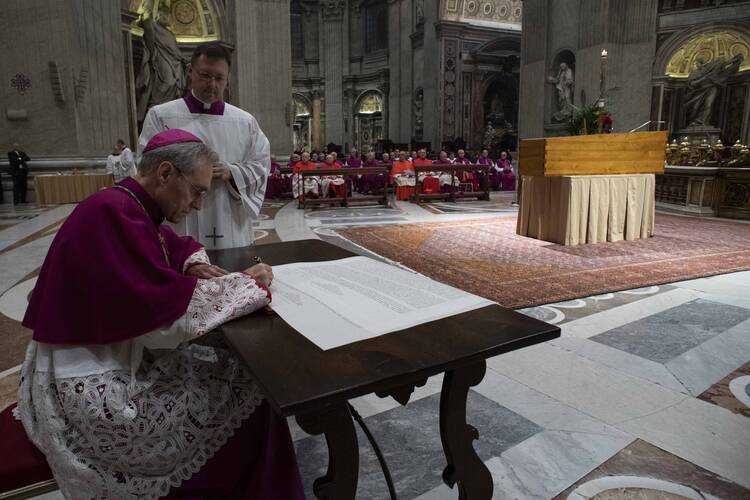 georg gänswein signs a paper in the foreground with the casket of benedict in the background with other priests and cardinals in the back