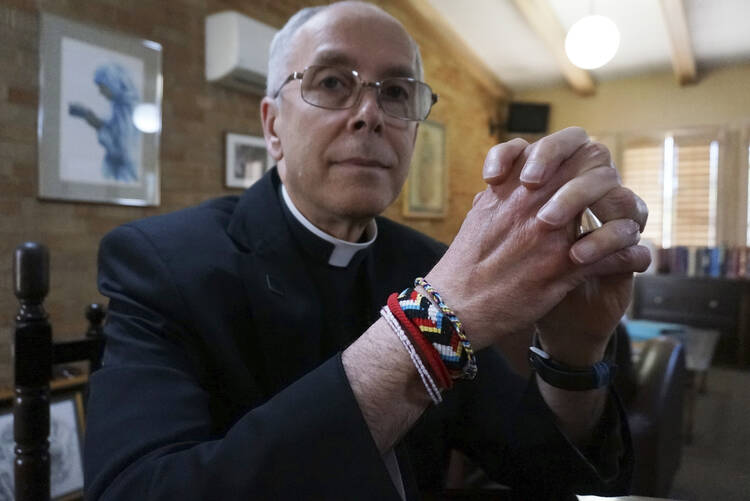 A man wearing black clerical clothing faces the camera, wearing a bracelet