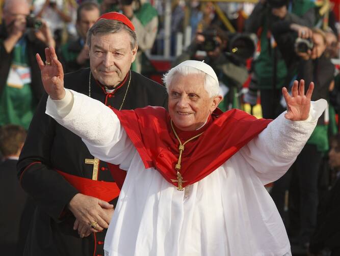 The former pope, wearing white and red, holds his arms out in greeting while Cardinal Pell stands behind him