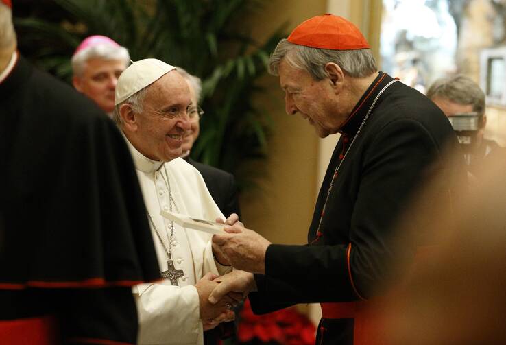 at left, pope francis smiles with his hand grabbing that of cardinal pell, who stands at right wearing black with a red cap