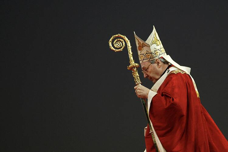 cardinal pell looks to the left wearing his miter and carrying his croiser while wearing fancy red vestments. a gray background is behind him