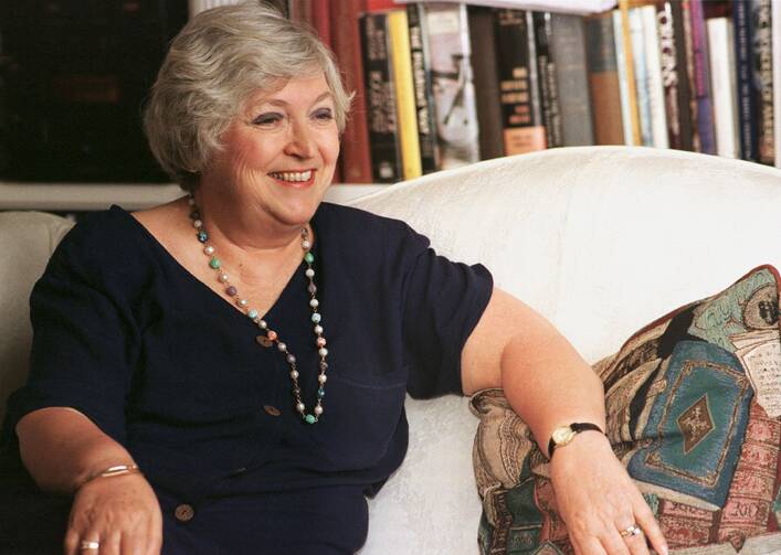 dolores leckey is seated wearing a black shirt and a necklace on a white couch with books behind her