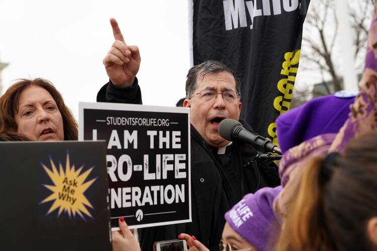 A priest speaks into a microphone in front of a sign that reads "I am the pro-life generation"