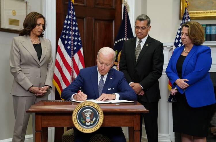 biden sits at a table wearing blue suit, presidential seal in front of him, kamala harris stands to his right, two others to his left.