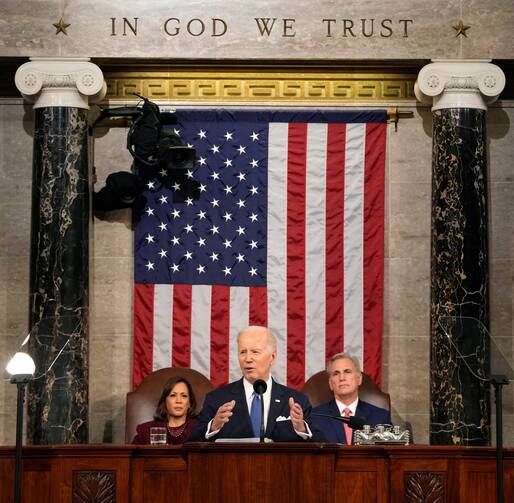 biden sits and gestures with kamala harris and kevin mccarthy behind him, the american flag hangs behind them and the words "in god we trust" are inscribed above the flag