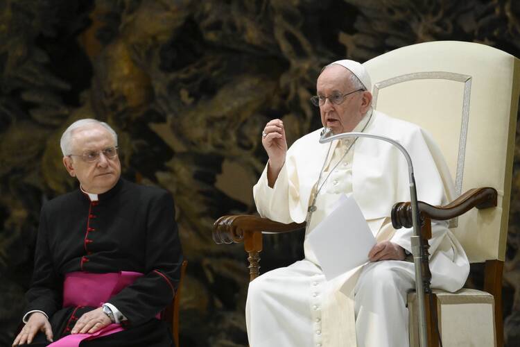 Pope Francis, seated and dressed in white papal robes, speaks into a microphone