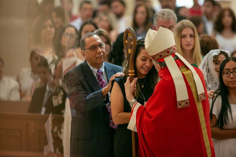 a bishop in a red chasuble and white hat makes the sign of the cross on the forehead of a 20-something woman being confirmed, many people are behind her