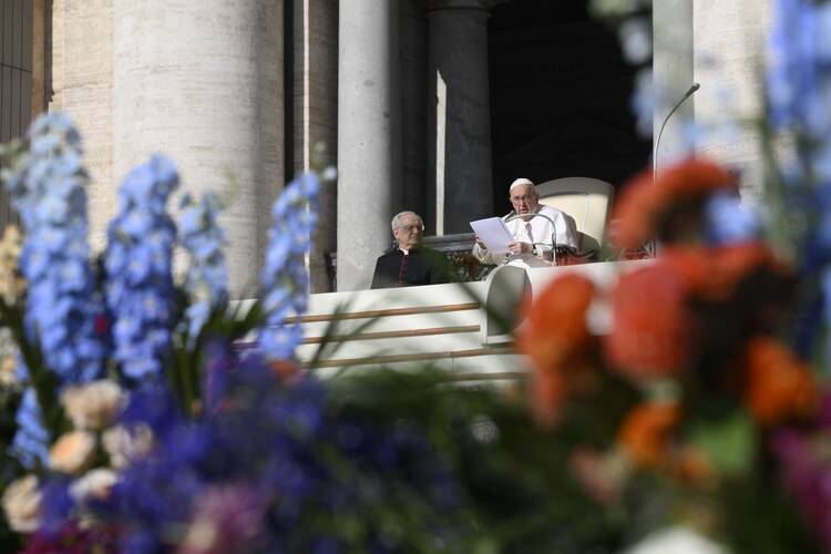 pope francis sits at his chair in the distances, some blue flowers are out of focus on the left front of the photo and some red ones on the right
