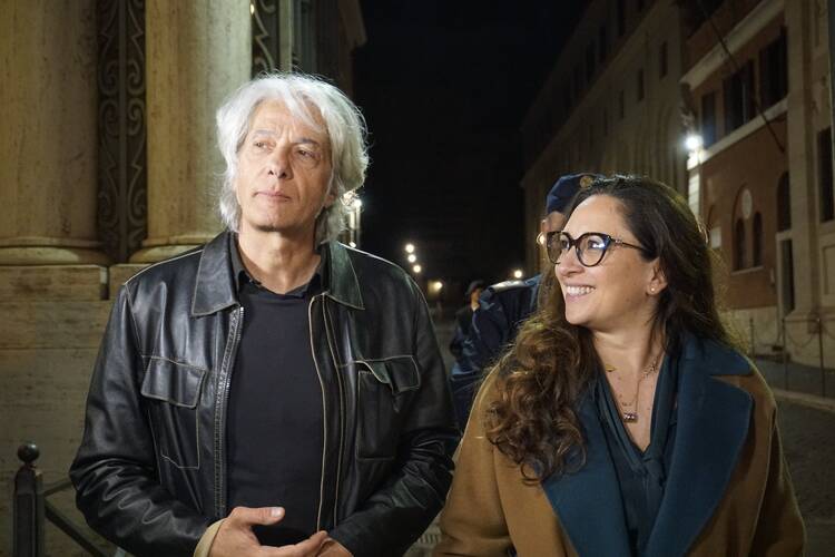 Pietro Orlandi in black leather jacket standing next to Laura Sgrò in dark Vatican street at night.