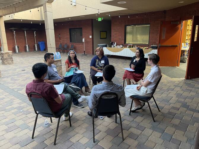 seven young people sit around in chairs talking to each other in a courtyard area inside a building