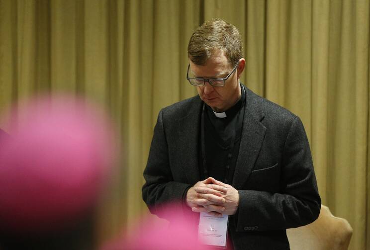 father hans zollner prays with head bowed in his clerics with a gold cloth-covered wall behind him; the purple caps of bishops or cardinals are blurred in front of him in the foreground