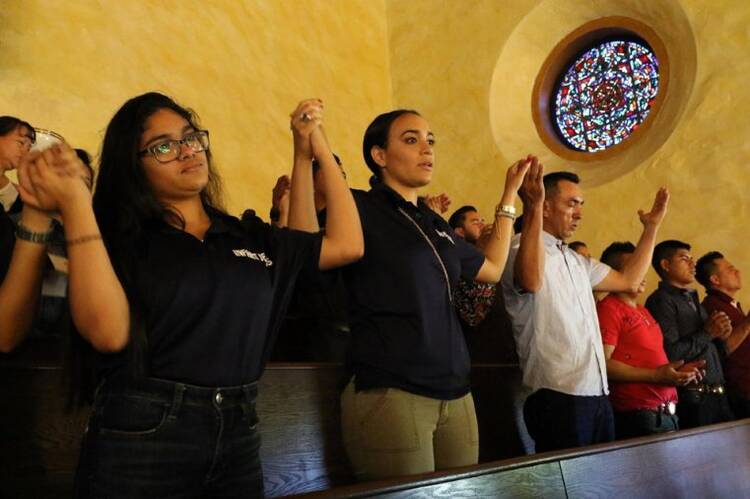 Latino Catholics in black and white shirts hold hands while reciting the Lord's Prayer during Mass inside a church under a circular stained glass window.
