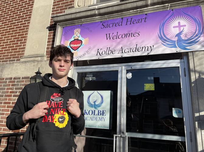 a student from kolbe academy stands at the front door of the school which has a sign with the school's name on it