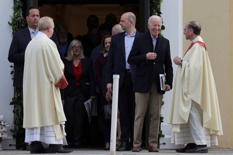 several people including joe biden walk out of a church building and are greeted by two priests in robes