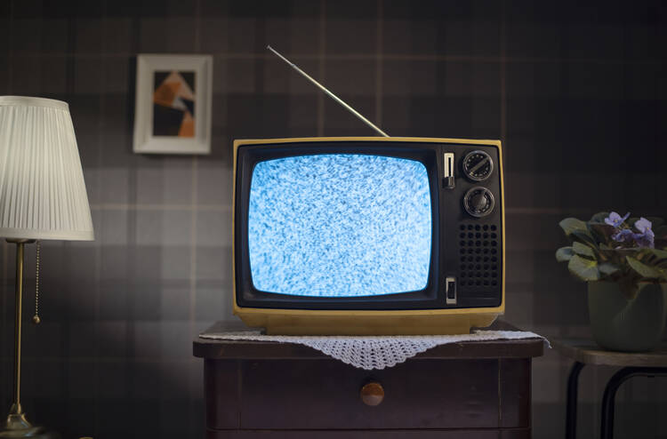 a static old television with antenna sits on a table with a doilie under it, a plaid wall is behind it. a very 1960s style image
