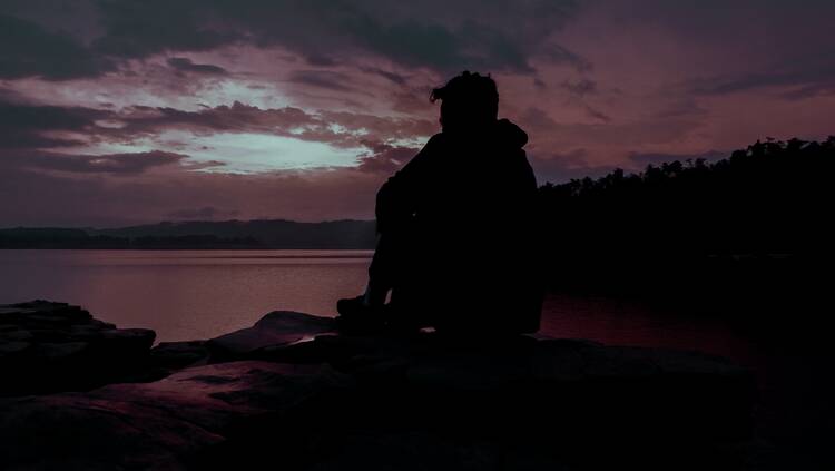 Silhouette of man sitting on rock near water during sunset