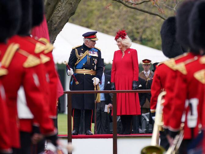 king charles in full regalia walks next to queen camilla who wears red with some british soldiers and guard in front of them