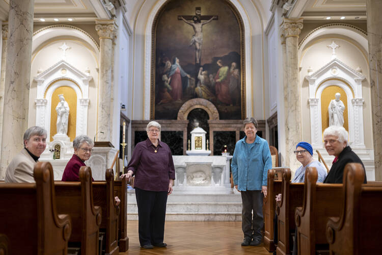 six sisters of charity stand in a chapel with the altar behind them, they are women in their 70s and wear regular clothes, not habits