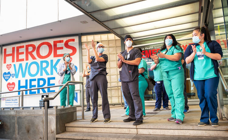 health care workers clapping outside a hospital wearing masks during covid, there is a sign behind them denoting them as heroes