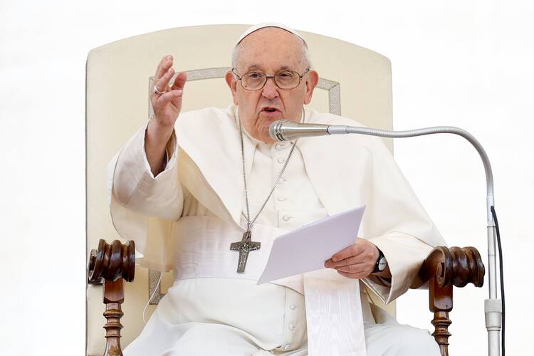 pope francis sits in his chair and gestures with his right hand while holding papers in his left