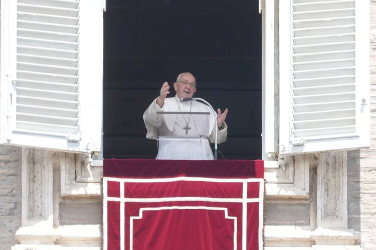 pope francis stands in a window gesturing and smiling with a red banner in front of him