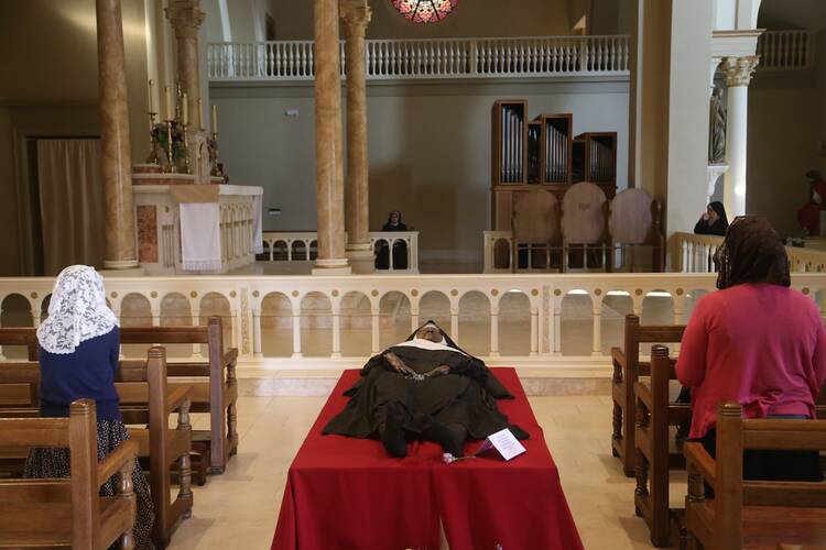 a nun's body is laid out in her habit atop a red covered platform, in the aisle of a chapel, you can see a few of the pews beside it