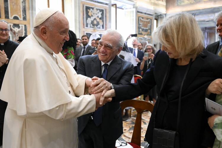 Pope Francis on the left shakes hands with Martin Scorsese in the center and Helen Morris Scorsese on the right in a daylight hall at the Vatican.