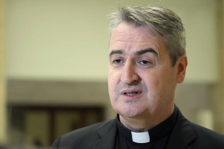 a priest wears a collar and talks in front of a beige background, he has gray hair and dark eyebrows