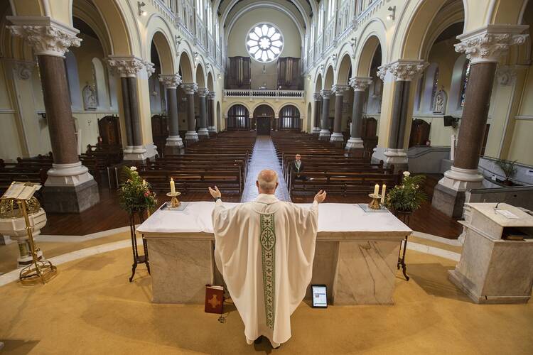 a priest stands behind an altar, the camera looks from the area where the tabernacle is, out onto the congregation. only one person sits in the pews in front of the priest, signifying the low number of people attending church.