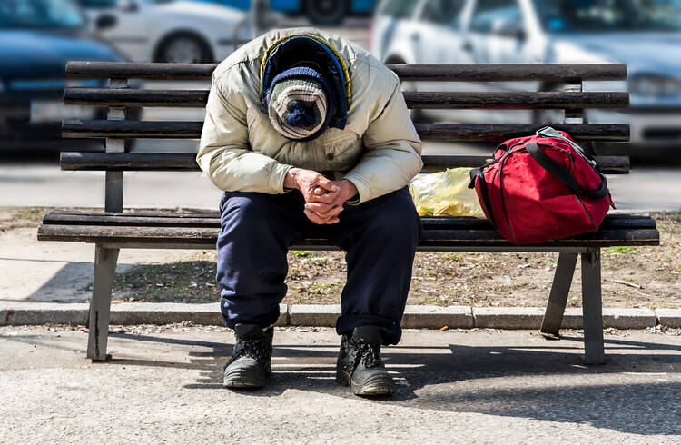 A man sits on a park bench with his head down and a cloth bag of his belongings next to him .(iStock/Srdjanns74)