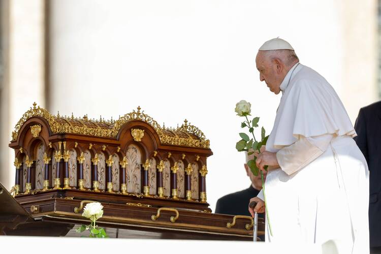 pope francis faces a red and gold decorated box with the relics of st therese of lisieux. he holds a white flower while facing it