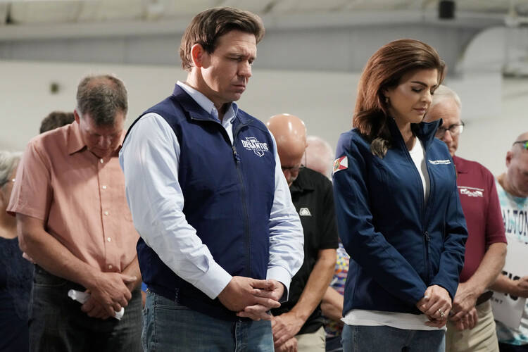 ron desantis and wife casey bow their heads during a prayer at a campaign event, they are standing at the front of a group of people, he wears a light blue button down with a navy vest atop