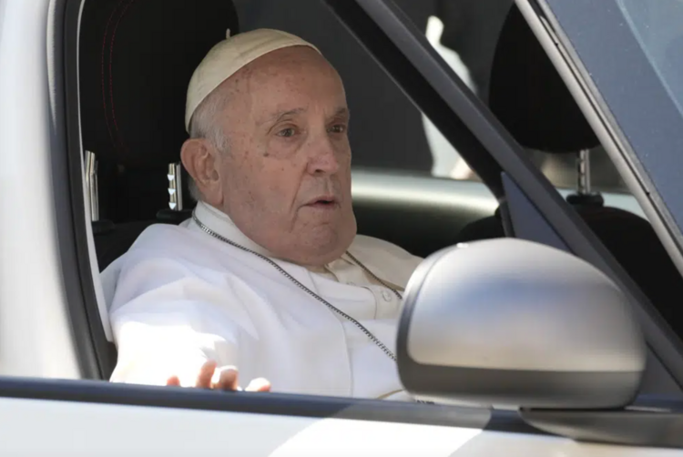 Pope Francis sits in the passenger seat of a silver car. 