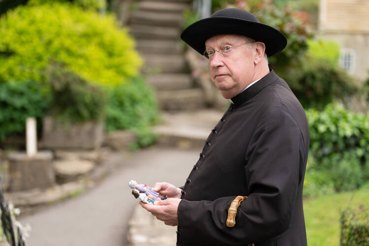 The actor Mark Williams in a scene from the series Father Brown, wearing a traditional black cassock, saturno hat and glasses
