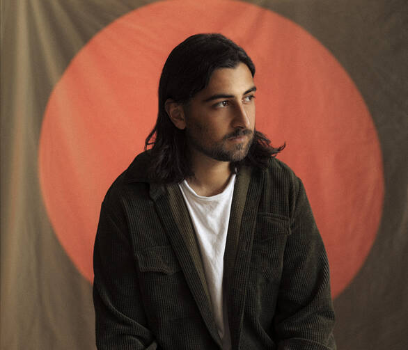 Noah Kahan wears a white shirt with a brown jacket in front of a brown and orange background.