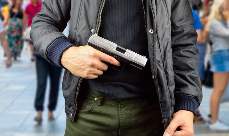 The torso of a man pulling a silver-and-black handgun from under his jacket, on a city street with several pedestrians visible behind him.