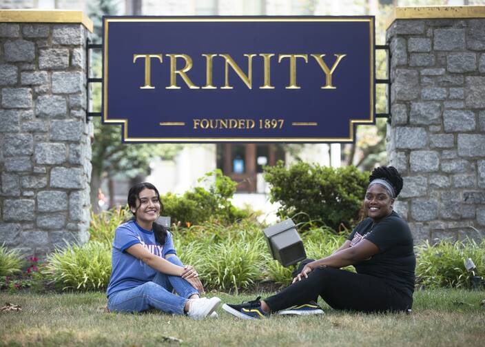 Two young women sit on the grass beneath the main entrance sign to Trinity Washington University, in Washington D.C., and smile toward the camera. (CNS photo/Tyler Orsburn)