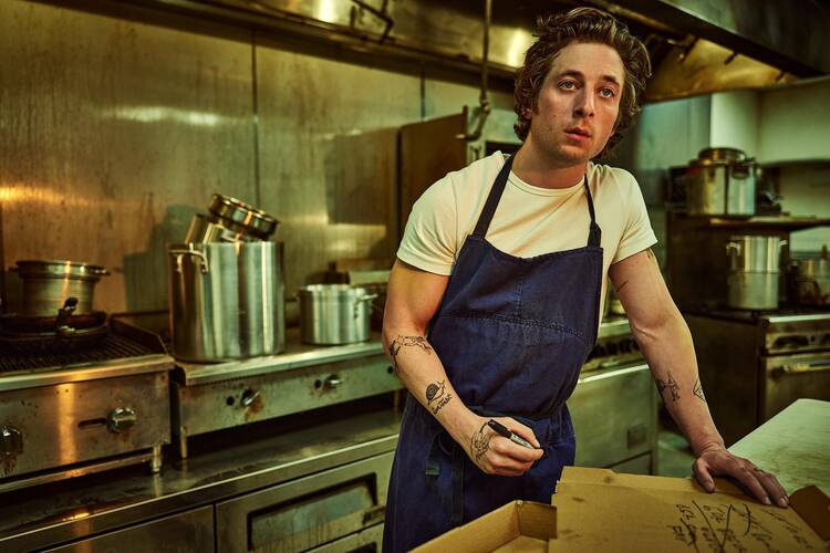 Character Carmy Berzatto stands in a kitchen with an apron on. 