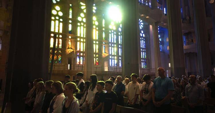 A crowd of people pray in the pews of Sagrada Familia basilica with stain glass windows behind them.