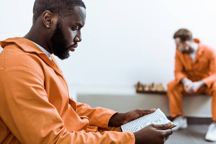 A young man in an orange prison uniform reads a book, while in the background another prisoner looks at a chessboard..