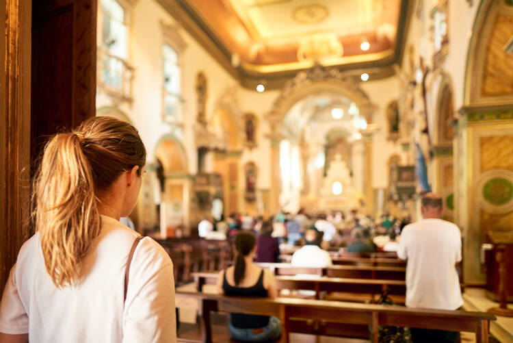 A woman with her back to the camera pauses before entering a Catholic church with several people sitting in the pews. 