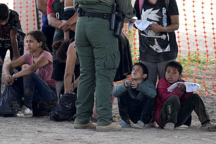 Young migrants who crossed into the U.S. from Mexico sit on the ground in Eagle Pass, Tex. The photo also shows a Border Patrol agent checking the papers of an adult migrant, but the heads of both are out of the frame. (AP Photo/Eric Gay)
