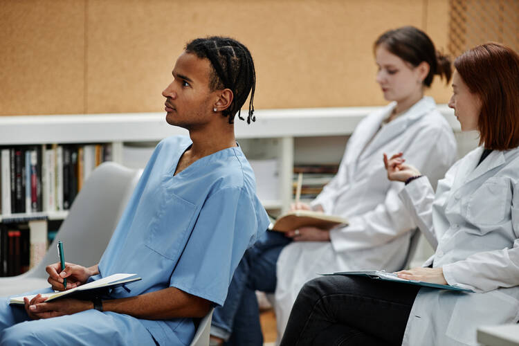 A group of students of different races wearing scrubs and white jackets and taking notes while sitting at classroom desks 