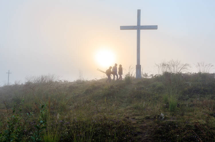 Three people are seen in silhouette approaching a large cross on a hill at sunrise (iStock/j-wildman)