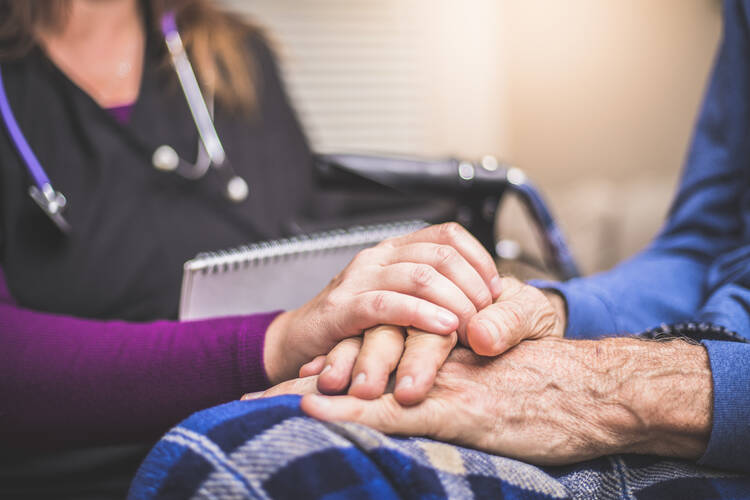 A hospice nurse (seated in a chair) cradles the hands of an elderly male hospice patient (sitting in a bed). (iStock)